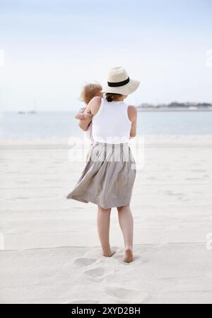 Vista posteriore della giovane madre vestita con cappello e canotta bianca con gonna grigia che tiene il bambino mentre si trova sulla sabbia bianca al mare durante il giorno estivo Foto Stock