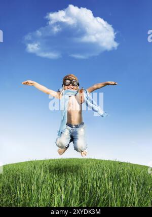 Capretto felice vestito come un pilota di saltare in campo verde contro il cielo blu. La vacanza estiva concept Foto Stock