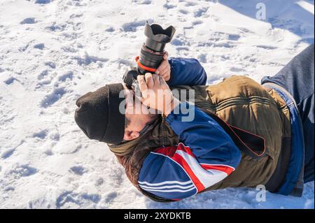 un uomo anziano con una macchina fotografica in mano, sdraiato sulla neve e fotografato un paesaggio invernale di montagna. Foto Stock