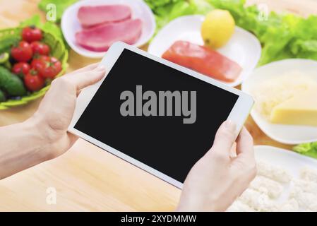 Vista dall'alto ravvicinata di mani femminili che tengono in mano un tablet computer con uno schermo vuoto circondato da cibo sano su un tavolo da cucina di casa. Pezzo da infilare Foto Stock