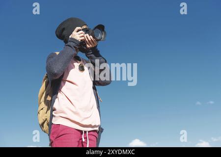 Ritratto di una ragazza hippy elegante con un cappello e uno zaino che la fotografa con una fotocamera reflex digitale all'aperto contro un cielo blu in una zona soleggiata Foto Stock