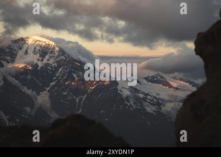 Un panorama al tramonto dell'elbrus e parte della cresta caucasica con nuvole arancioni e un ghiacciaio incrinato sul fondo Foto Stock