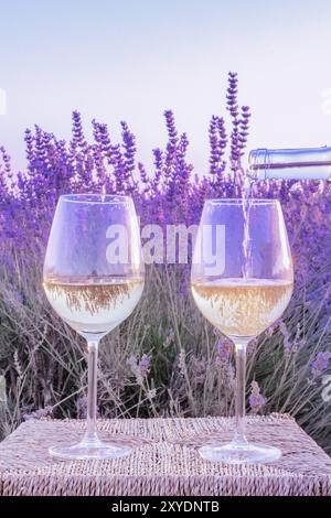 Vino di lavanda. Vino bianco versata da una bottiglia in bicchieri contro un campo di lavanda sfondo, immagine dai toni Foto Stock