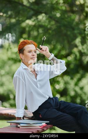 Insegnante dai capelli rossi in camicia bianca, pantaloni neri, seduta sulla panchina persa nel pensiero con la penna Foto Stock