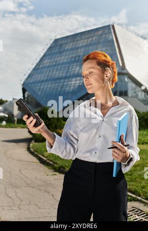 Una donna con i capelli rossi corti attraversa il campus, tenendo in mano un telefono e una cartella blu. Foto Stock