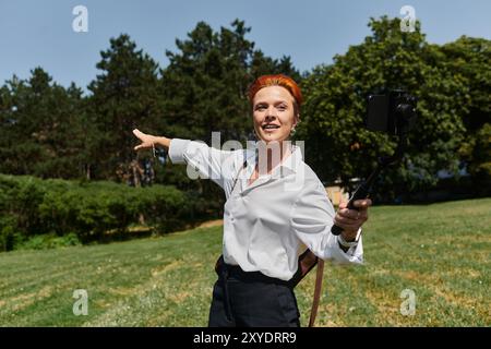 Una donna con i capelli rossi sorride alla telecamera mentre scatta un selfie in un campus soleggiato. Foto Stock