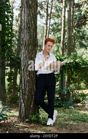 Un insegnante si appoggia contro un albero, leggendo un libro nel bosco. Foto Stock