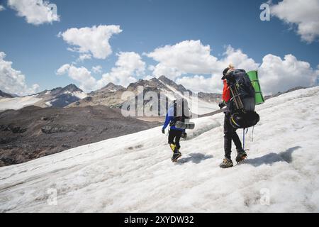 Due turisti, un uomo e una donna con zaini e ramponi ai piedi, camminano lungo il ghiacciaio sullo sfondo delle montagne del cielo Foto Stock