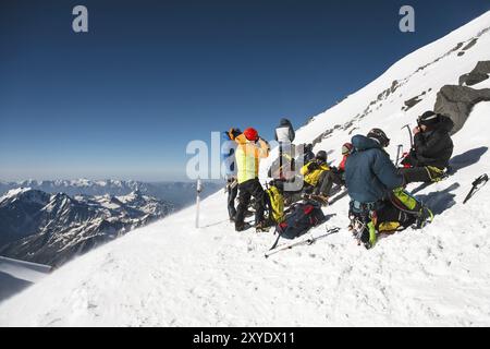 Alpinisti professionisti completamente attrezzati in sosta siedono su una pista innevata con il sole. Il concetto di ricreazione collettiva in montagna Foto Stock