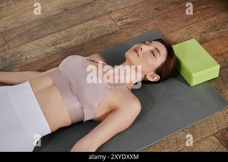 Una donna asiatica, che indossa una copertura, giace su un tappetino da yoga nel suo appartamento, gli occhi chiusi, con un blocco verde da yoga sotto la testa. Foto Stock