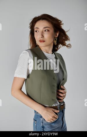 Un'elegante donna asiatica si posa su uno sfondo grigio, indossando un gilet verde su una t-shirt bianca e un jeans. Foto Stock