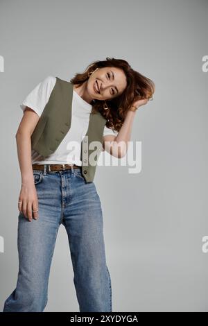 Un'elegante donna asiatica che indossa un gilet verde, una t-shirt bianca e jeans si posa su uno sfondo grigio. Foto Stock