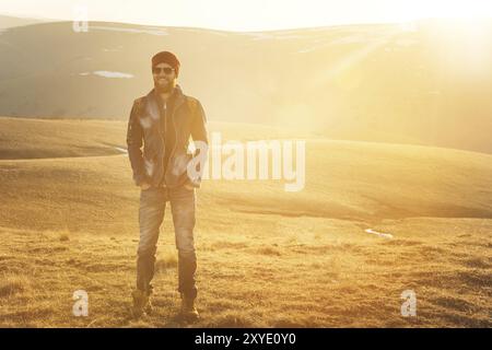 Ritratto alla moda di un giovane hipster barbuto che indossa occhiali da sole, uno zaino e un cappello su uno sfondo con copyspase sulle montagne al tramonto, Un co Foto Stock