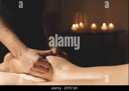 Primo piano di mani maschili che fanno un massaggio polpaccio di gambe femminili in una stanza buia con candele sullo sfondo. Trattamenti cosmetici e spa Foto Stock