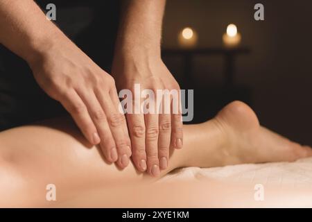 Primo piano di mani maschili che fanno un massaggio polpaccio di gambe femminili in una stanza buia con candele sullo sfondo. Trattamenti cosmetici e spa Foto Stock