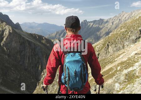 Un uomo barbuto con occhiali da sole e un berretto con uno zaino si staglia sulla cima di una roccia e guarda verso una valle rocciosa in alto tra le montagne. Il concetto di tour Foto Stock