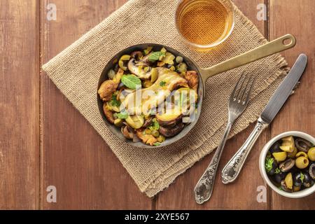 Rustico di funghi e olive rosolare in una ghisa pan, con un bicchiere di vino bianco, girato da sopra al buio su un sfondo rustico con un posto per il testo Foto Stock