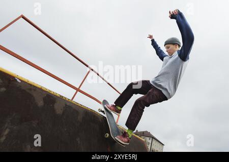 Uno skateboarder adolescente con un cappello fa un trick di Rocks su una rampa in uno skate Park contro un cielo nuvoloso e una zona notte. Il concetto di stile urbano in sp Foto Stock