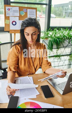 Una donna con i capelli castani scuri si siede alla sua scrivania, rivedendo documenti e documenti. Foto Stock