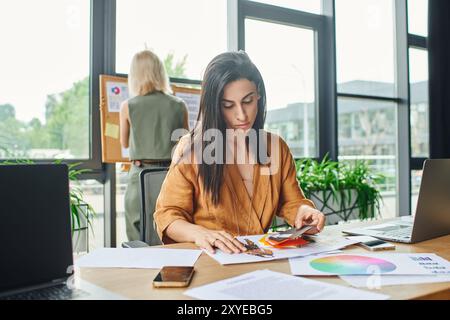 Una donna con una camicetta gialla brillante esamina una tavolozza di colori, perduta nel pensiero mentre lavora in un ambiente di ufficio contemporaneo. Foto Stock