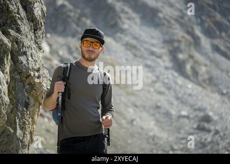 Porter di un elegante viaggiatore hipster con una barba e uno zaino in occhiali da sole e un berretto con pali da trekking si erge su una roccia sullo sfondo di Foto Stock