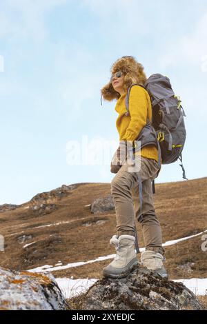 La ragazza con gli occhiali da sole e un grande cappello di pelliccia del nord con uno zaino sulla schiena è in piedi su una roccia e guarda verso le scogliere che si nascondono dentro Foto Stock