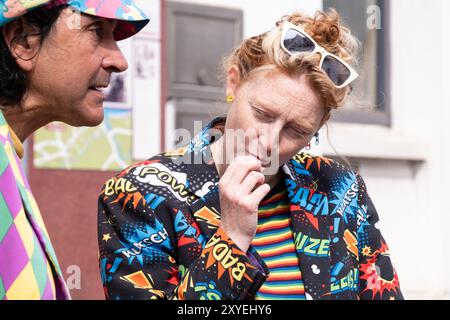 Donna con occhiali da sole in capelli rossi vestiti con giacca elegante, riferimenti comici Old School accanto all'uomo in giacca brillante. Ballycastle, Regno Unito - 26 agosto 2024. Foto Stock