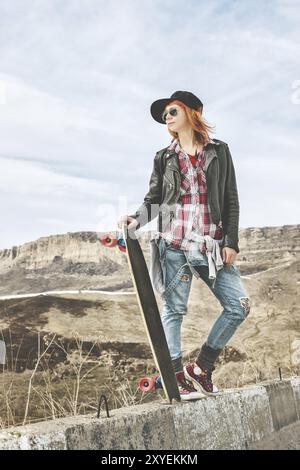 Eleganti tute ap e denim da donna dai capelli rossi in piedi su un dosso in autostrada in montagna con un longboard Foto Stock