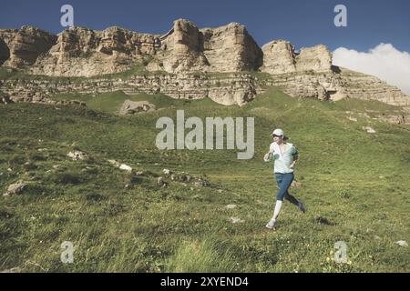 Attraente ragazza sportiva con un berretto e cuffie che fa jogging in un luogo pittoresco giù per la collina vicino alle rocce al tramonto. Allenati all'aperto Foto Stock