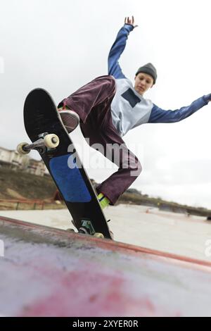 Uno skateboarder adolescente con un cappello fa un trick di Rocks su una rampa in uno skate Park contro un cielo nuvoloso e una zona notte. Il concetto di stile urbano in sp Foto Stock