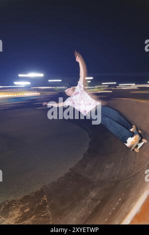 Un giovane skateboarder cavalca a velocità laterale lungo la rampa, spargendo le mani ai lati. Riprese notturne con lunga esposizione a velocità notturna in una ska Foto Stock