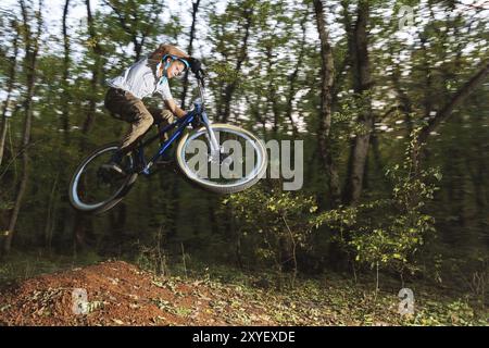 Un giovane pilota con un casco vola su una bicicletta dopo essere saltato da un kicker alto su una pista ciclabile nella foresta Foto Stock