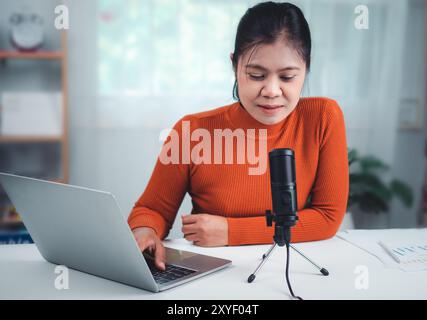 Donna che parla con il microfono sul supporto, conduce la comunicazione online, discute di un argomento, guarda lo schermo del notebook, impegnata in streaming, vigilia di riunione virtuale Foto Stock