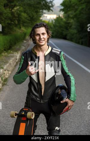 Un giovane uomo con un abito in pelle e capelli lunghi sta sull'asfalto con un casco tra le mani e il suo longboard sullo sfondo del verde e. Foto Stock