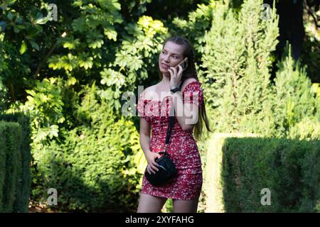 Una donna gioiosa dai capelli intrecciati, vestita con un abito estivo floreale rosso, gode di una piacevole conversazione sul suo smartphone circondato dal verde. Foto Stock