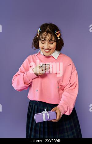 Una donna sorride brillantemente mentre guarda il suo telefono, tenendo un regalo nell'altra mano. Foto Stock