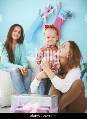 Ritratto di una madre con una bambina e una figlia adolescente che si avvicinano e si abbracciano a casa vicino all'albero di Natale e sono felici e gioiosi Foto Stock