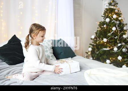 Incantevole piccola ragazza seduta su di un letto per bambino in background di albero di Natale nel luminoso interno della casa Foto Stock