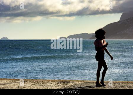 Ragazza camminare davanti al mare e guardando il telefono cellulare durante il pomeriggio Foto Stock