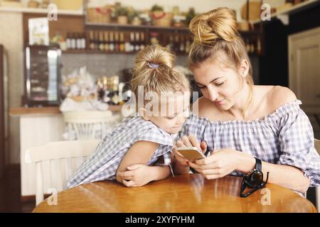 Mamma e bambina felici che cenano e usano lo smartphone al ristorante. Famiglia, genitorialità, concetto tecnologico Foto Stock