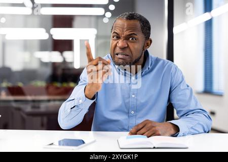 Uomo d'affari frustrato che indossa una camicia blu che mostra rabbia durante la videochiamata, gestendo con enfasi con le dita. Espressione intensa durante le riunioni virtuali in un moderno ambiente di ufficio Foto Stock