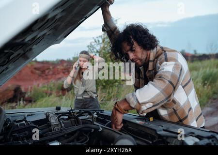 Giovane uomo con capelli ondulati scuri che si piega sul cofano aperto del pick-up e controlla il motore contro la sua ragazza chiamando il servizio di riparazione Foto Stock