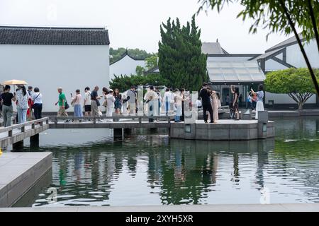 Suzhou, Cina - 12 giugno 2024: Un gruppo di persone si trova su un ponte e una piattaforma di pietra che si affaccia su uno stagno con riflessi dell'architettura e della t Foto Stock