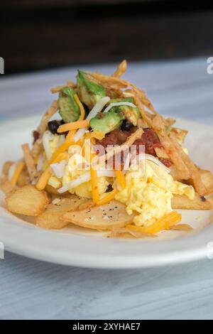 Hash per la colazione su un piatto bianco con uova, patate, formaggio, salsa, avocado, e strisce di tortilla Foto Stock