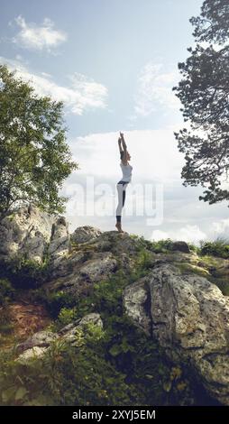 Vista laterale della giovane femmina slim attiva nello sport praticante yoga e salutare il sole con le mani sollevate mentre si sta in piedi sulla pittoresca cima di un moun roccioso Foto Stock