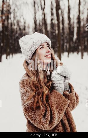 Bella giovane donna in neve fantasia bosco invernale. Ragazza che indossa morbidi guanti, cappuccio e cappotto. Foresta di Natale, alberi su sfondo sfocato. Incrociato Foto Stock