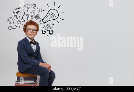 Simpatico bambino in agguato con lampadina e punti interrogativi seduti su una pila di libri su sfondo bianco. Educazione e concetto di idea Foto Stock