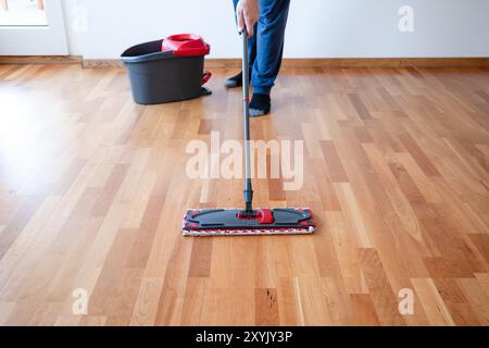 Uomo irriconoscibile che usa un mocio per la pulizia su un pavimento in piastrelle di legno dell'appartamento o pavimento in parquet. Scatto grandangolare, messa a fuoco selettiva, luminosità naturale della finestra Foto Stock