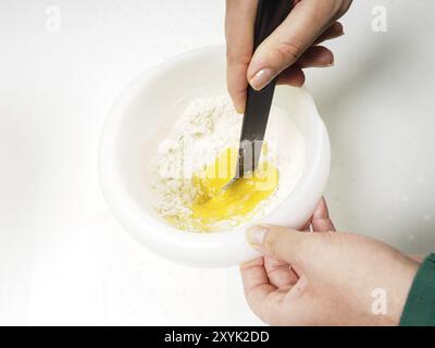 Donna che mescola la farina con tuorlo d'uovo e burro fuso con una spatola nera in un recipiente di plastica Foto Stock