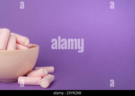 una vista dettagliata di una ciotola rosa piena di soffici marshmallow. Una piccola pila di marshmallow è posizionata a sinistra, il tutto contro un ricco viola Foto Stock